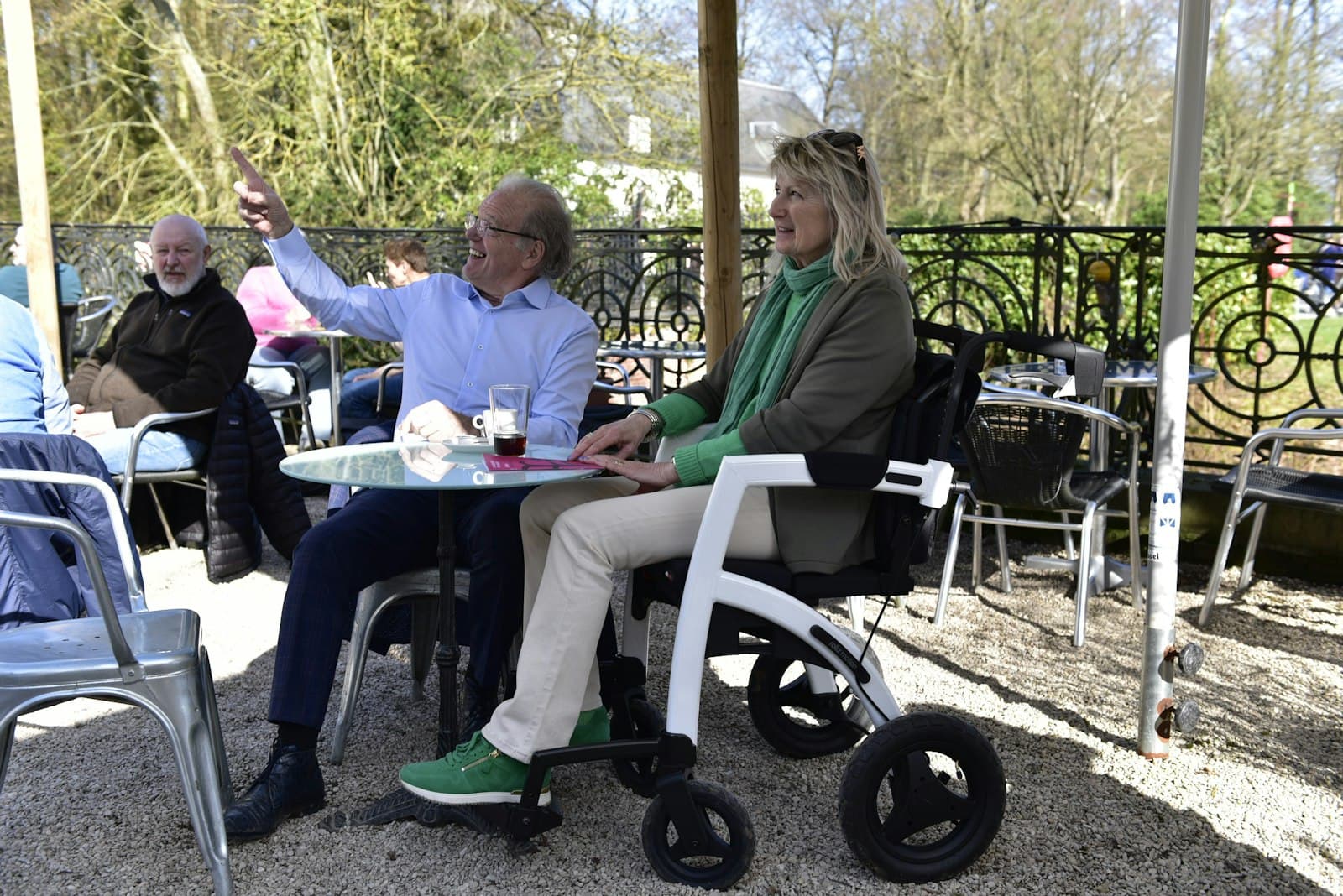 Una pareja mayor charla en la terraza de una cafetería, ella sentada en una silla de ruedas ligera junto a la mesa