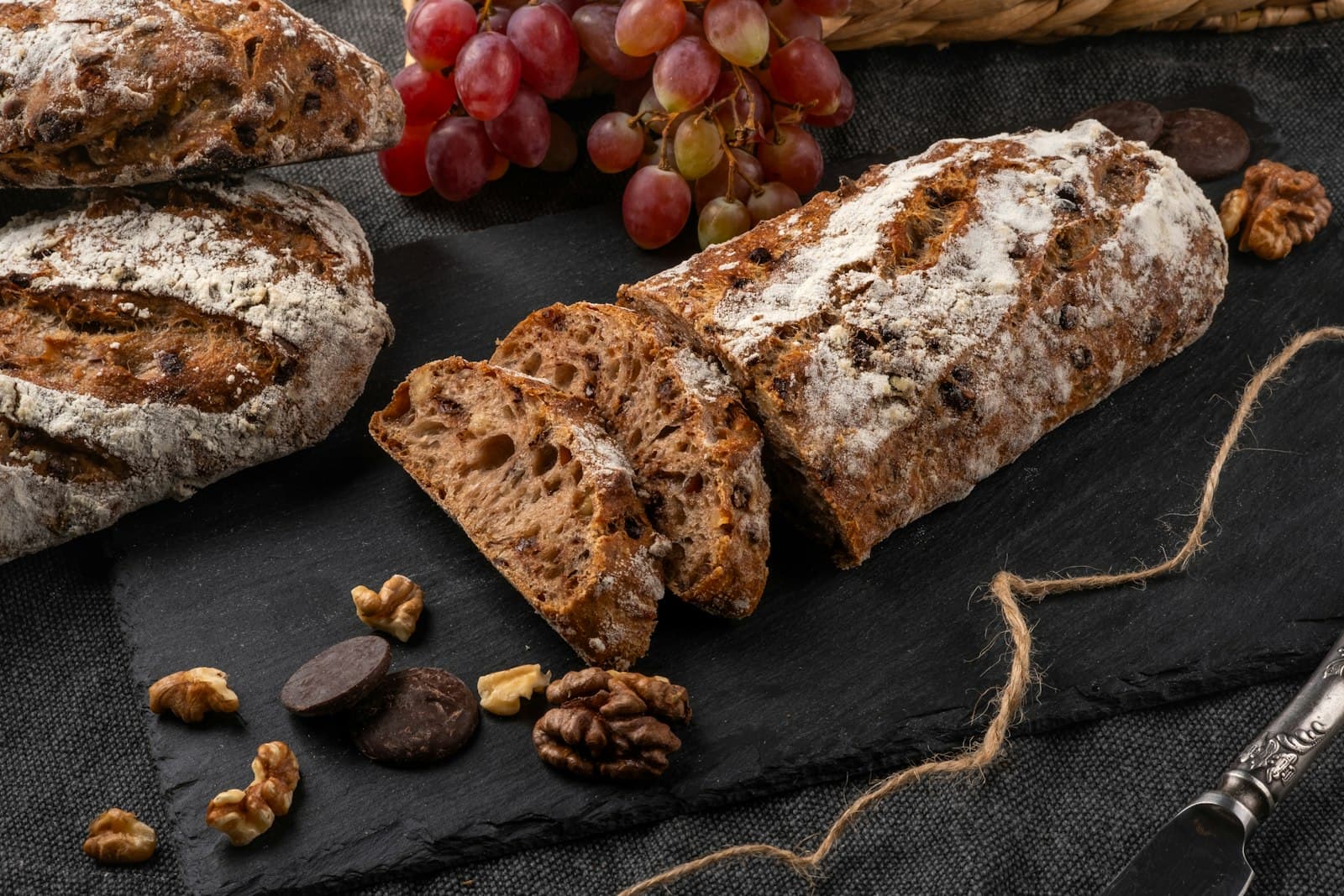 Pan de nueces cortado sobre una pizarra con uvas, nueces y chocolate alrededor