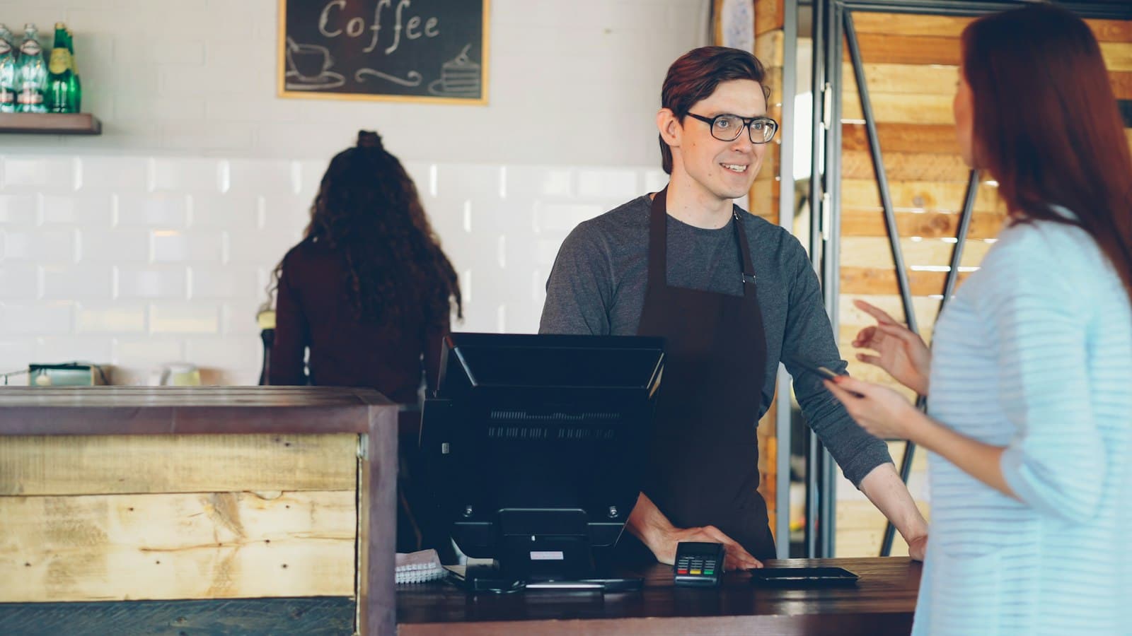 Una clienta con el móvil en la mano frente al mostrador de una cafetería, donde un camarero con delantal la atiende junto a la caja y el datáfono
