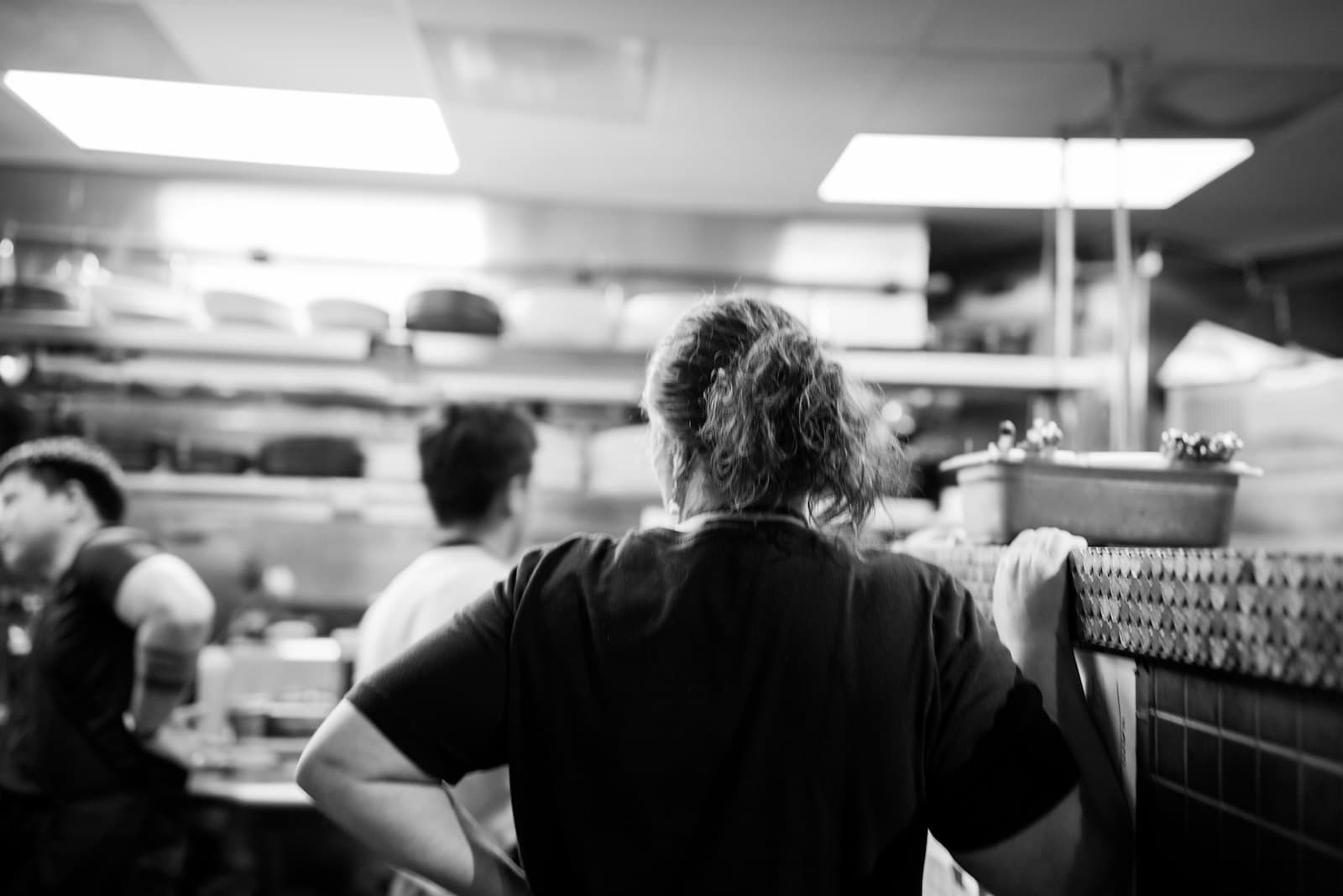 Una cocinera de espaldas en la cocina de un restaurante, con dos compañeros trabajando al fondo, en blanco y negro