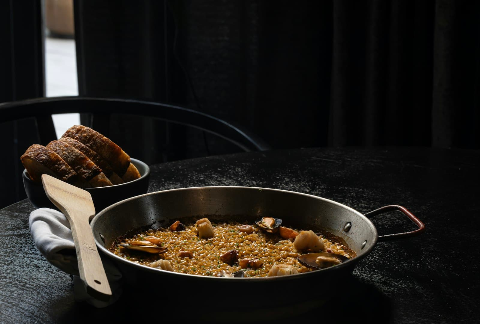 Paella con mejillones y un cuenco con rebanadas de pan sobre la mesa oscura de un restaurante