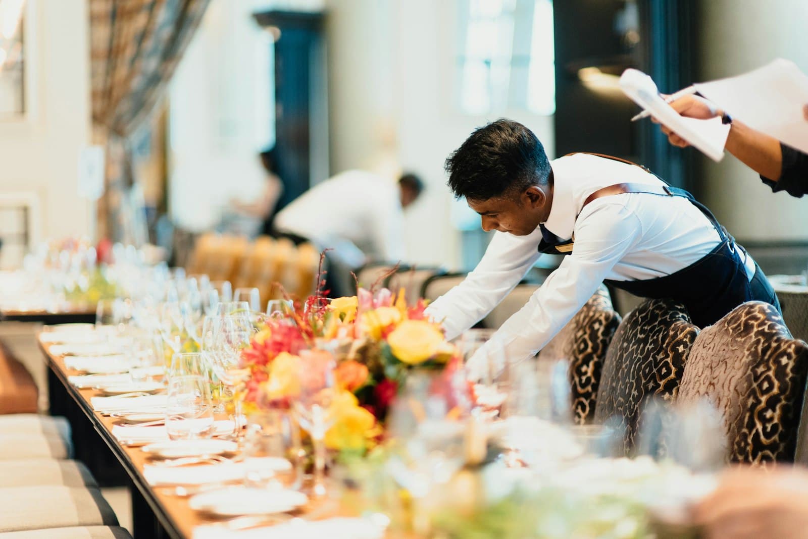 Camarero con delantal coloca la vajilla en una mesa larga de banquete decorada con flores