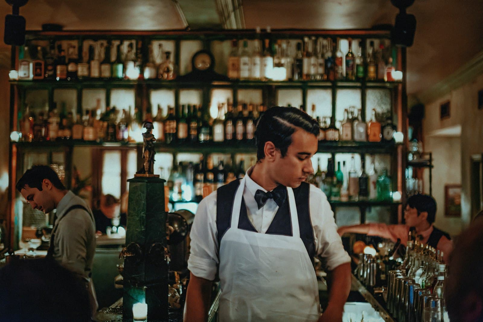 Camarero con delantal blanco y pajarita detrás de la barra de un bar, con otros compañeros trabajando al fondo