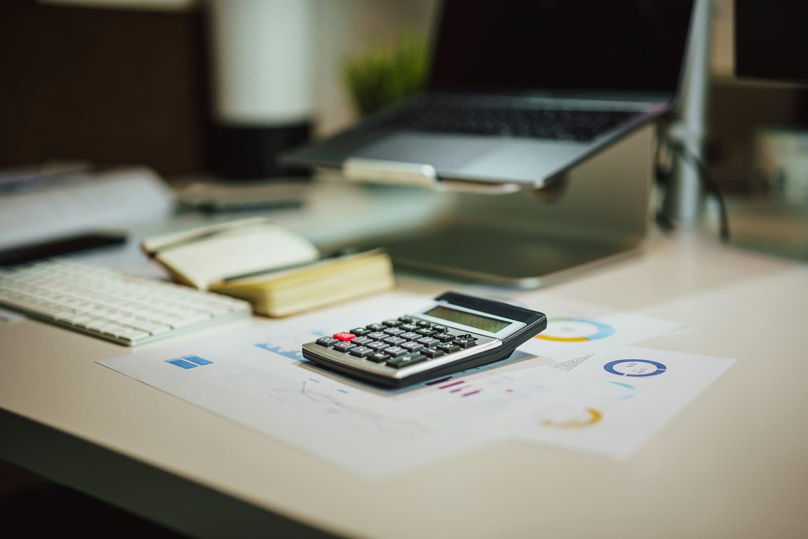 Calculadora sobre papeles con gráficos en una mesa de oficina, con un portátil al fondo