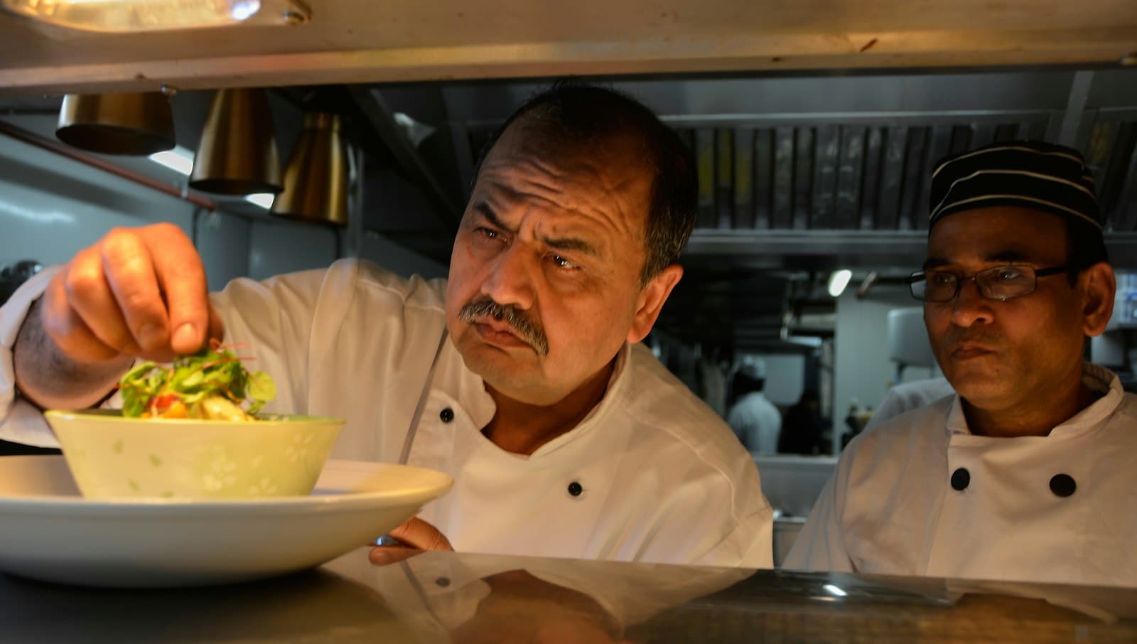 Cocinero revisando un plato en el pase mientras un compañero observa en la cocina