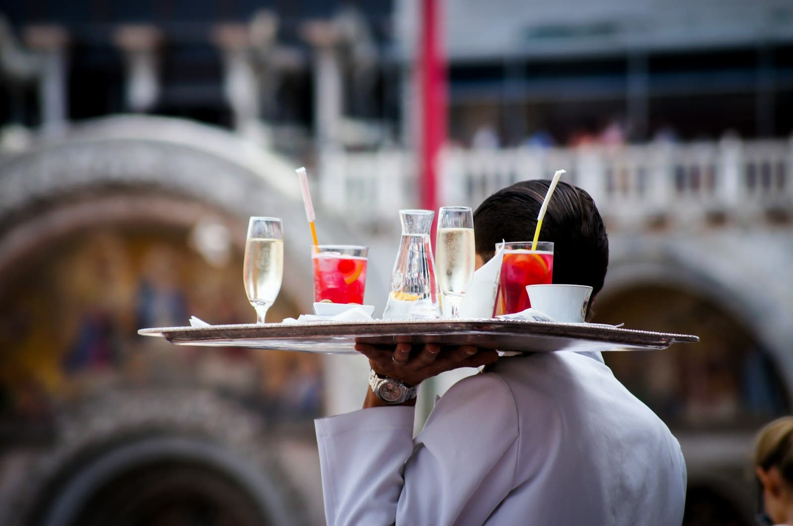 Camarero de chaqueta blanca lleva en alto una bandeja con copas de cava y refrescos frente a una fachada histórica