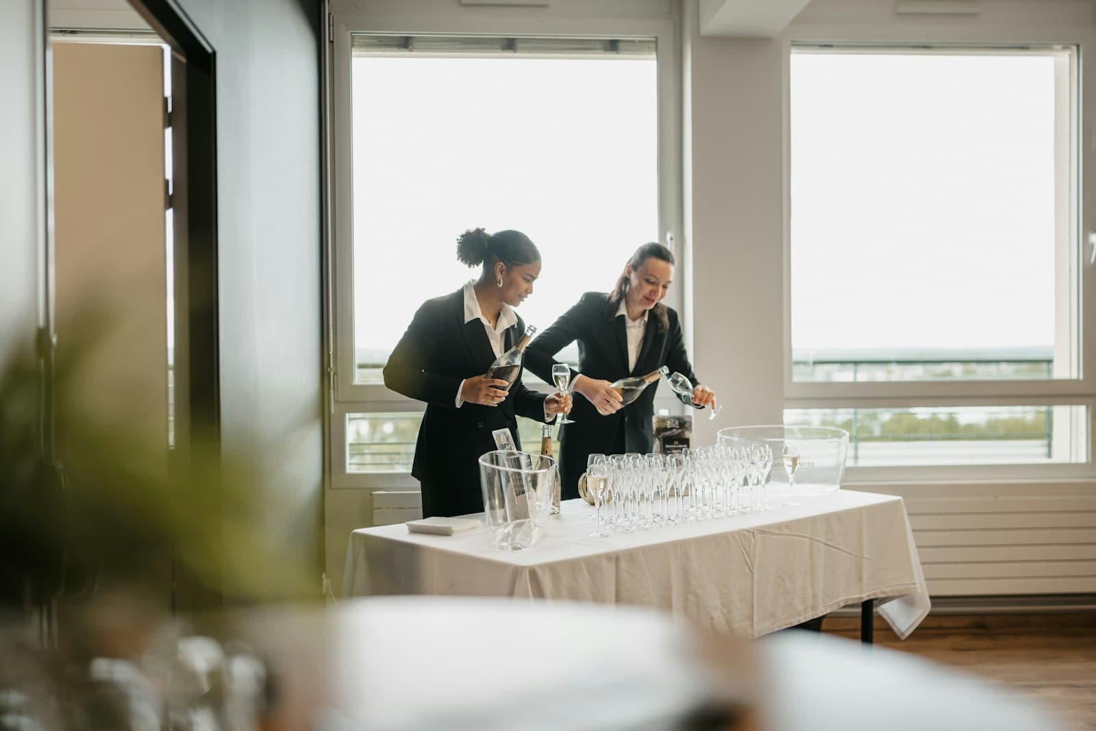 Dos camareras con traje oscuro sirven copas en una mesa con mantel blanco antes de un evento