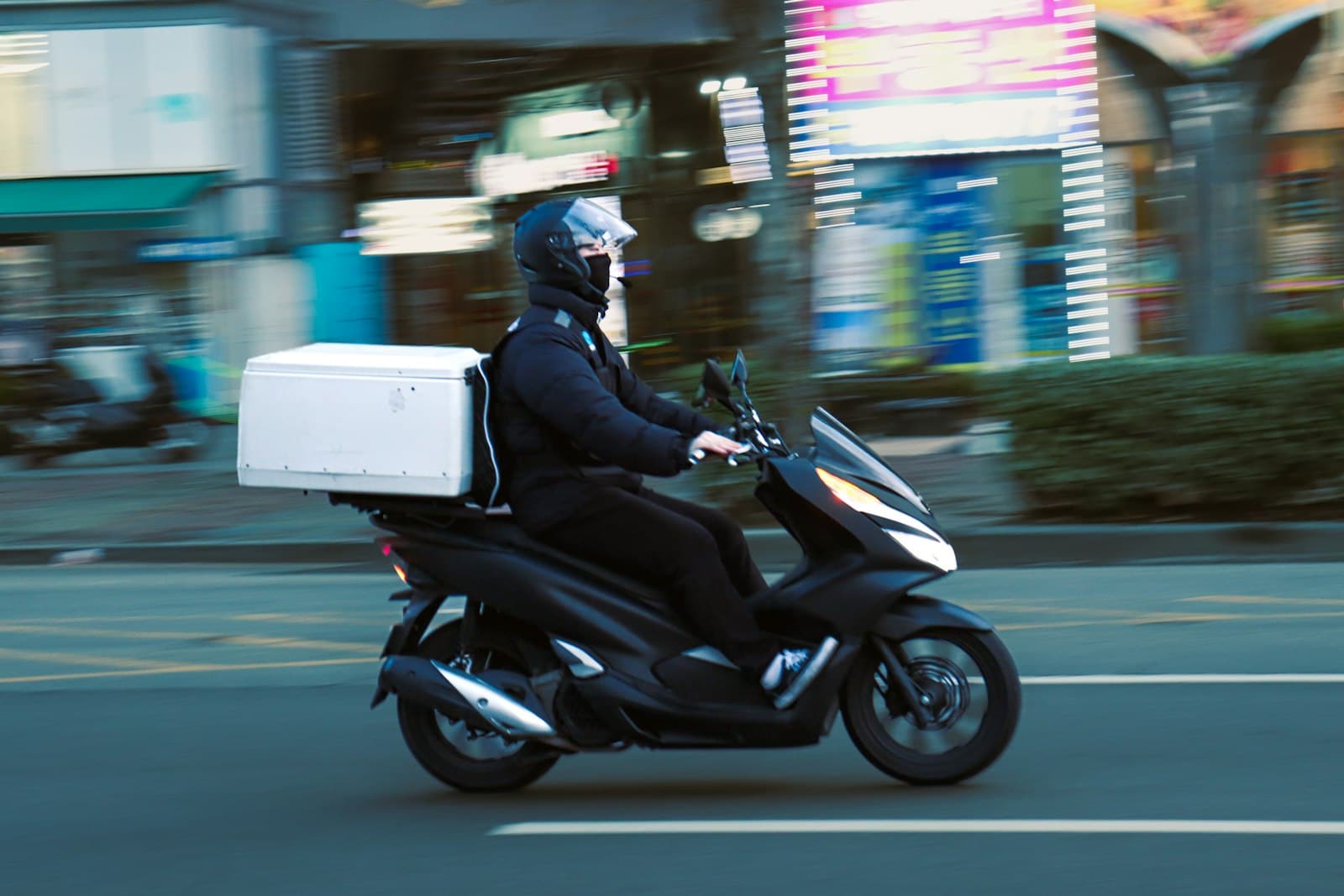 Repartidor con casco y una caja blanca de reparto en una moto negra, por una calle de ciudad
