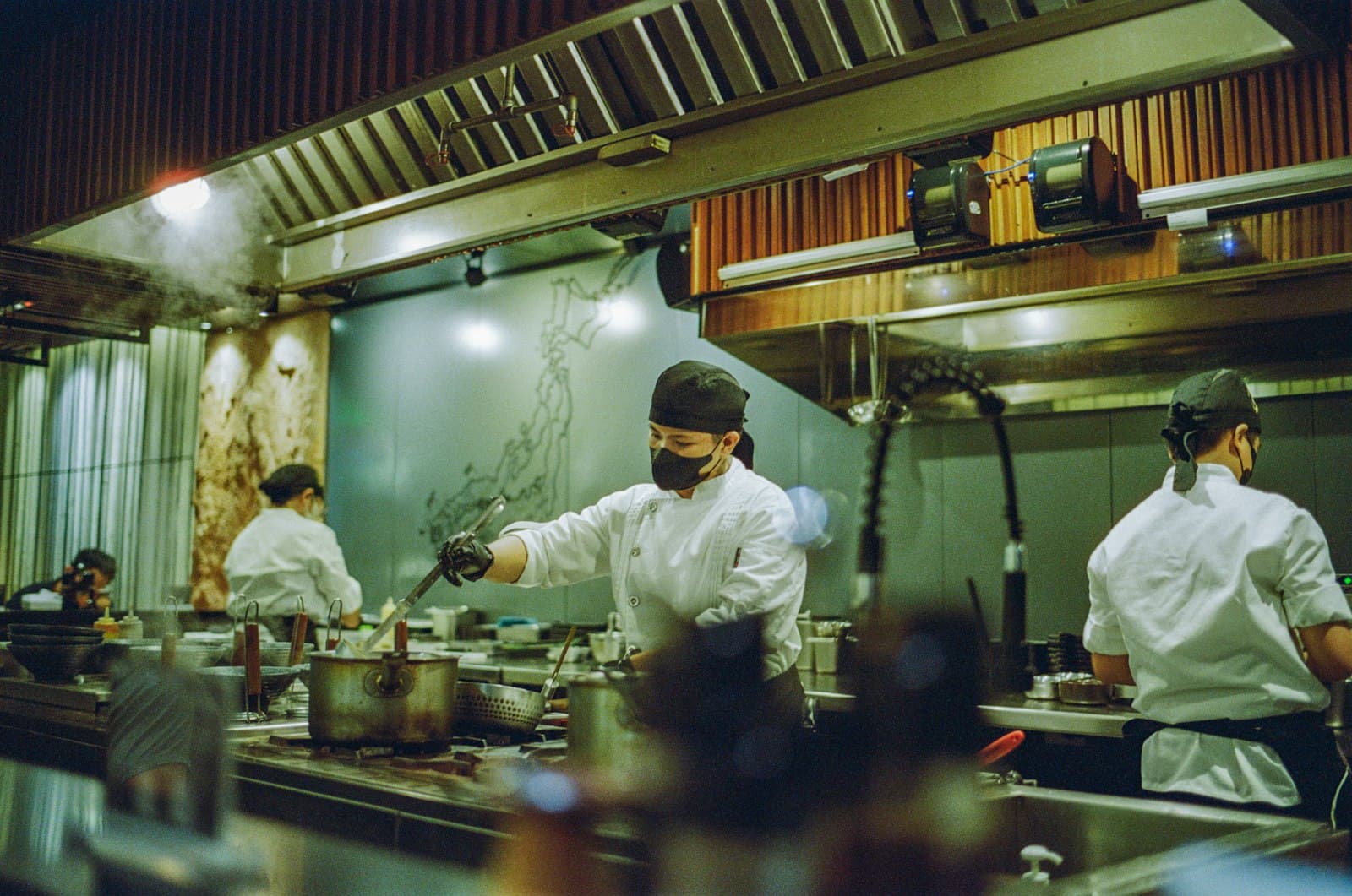 Cocineros con mascarilla trabajando en una cocina abierta con vapor e impresoras de comandas sobre la línea