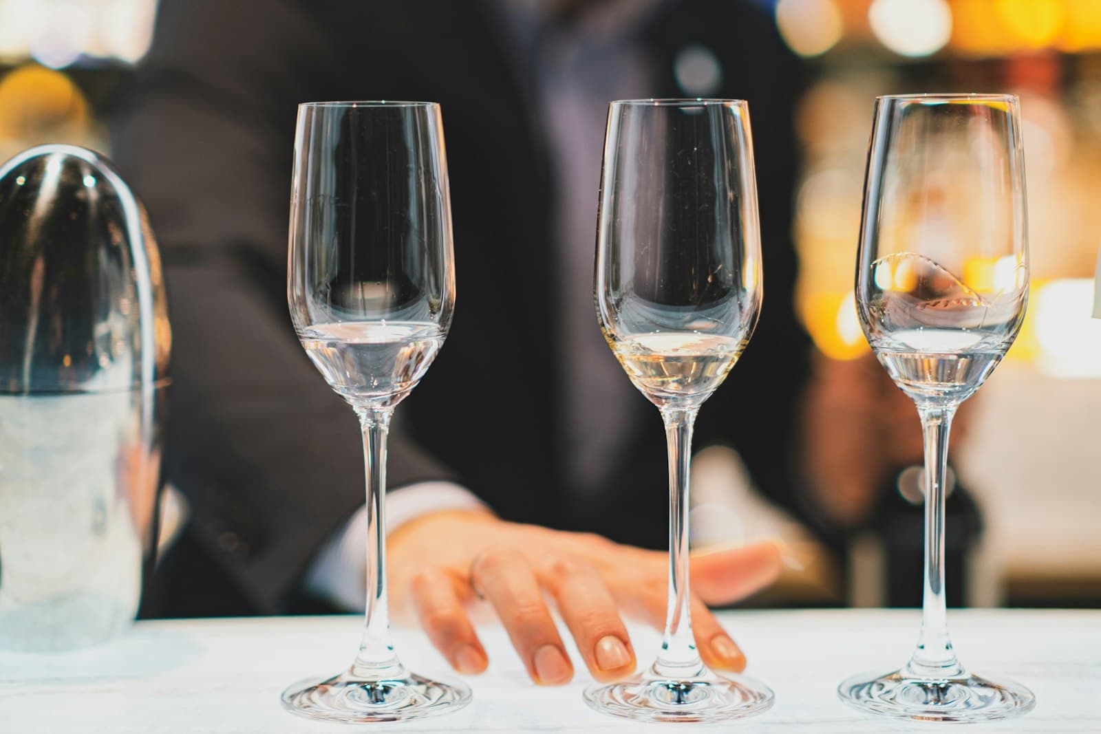Tres copas sobre la barra de un bar, con la mano del camarero detrás y luces cálidas al fondo