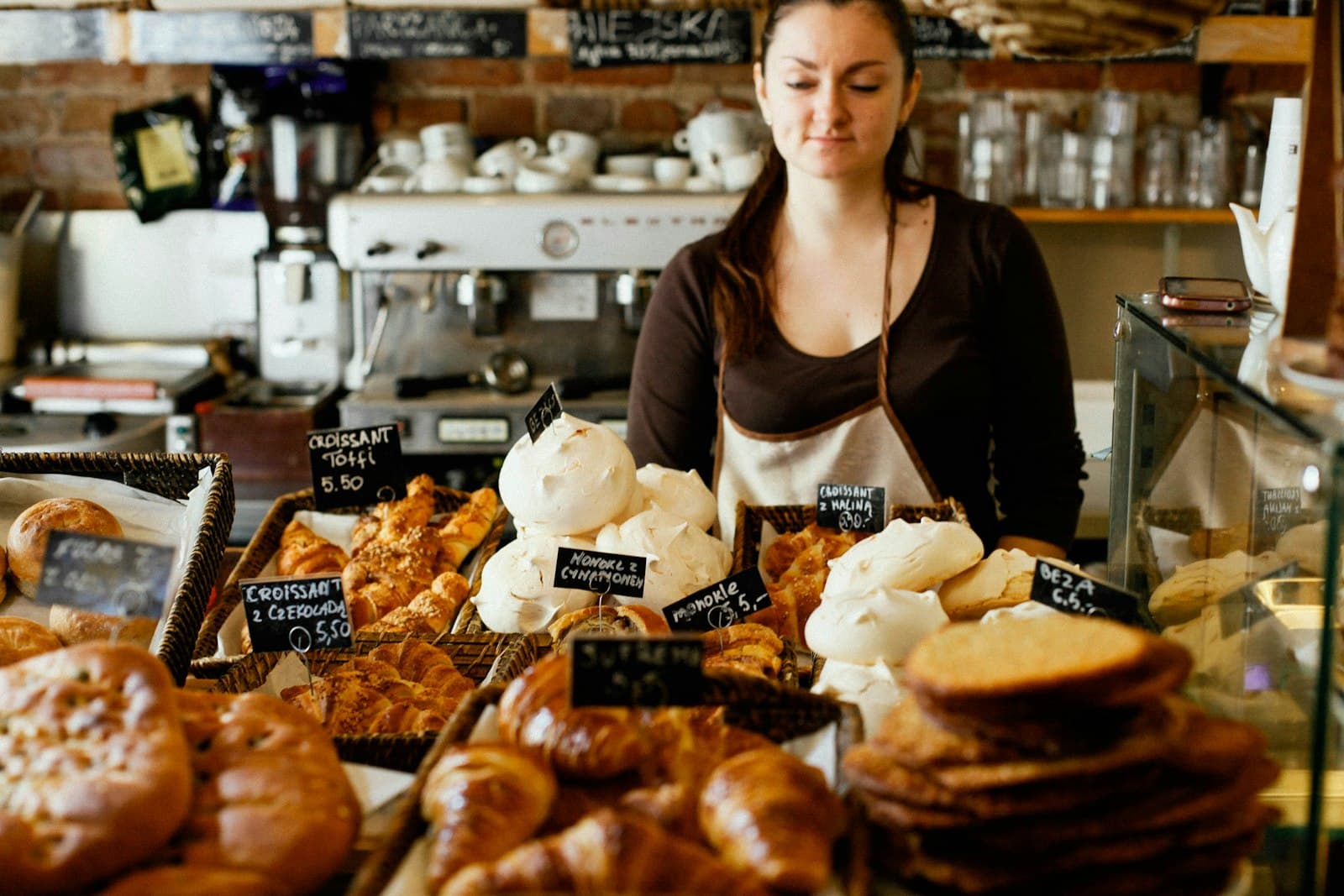 Dependienta detrás del mostrador de una cafetería panadería con cruasanes, merengues y precios en pizarras