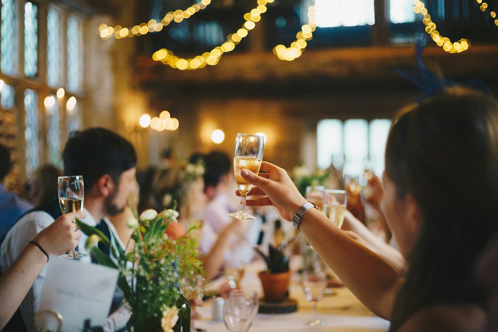 Comensales brindando con copas de cava en una mesa larga de celebración, bajo guirnaldas de luces