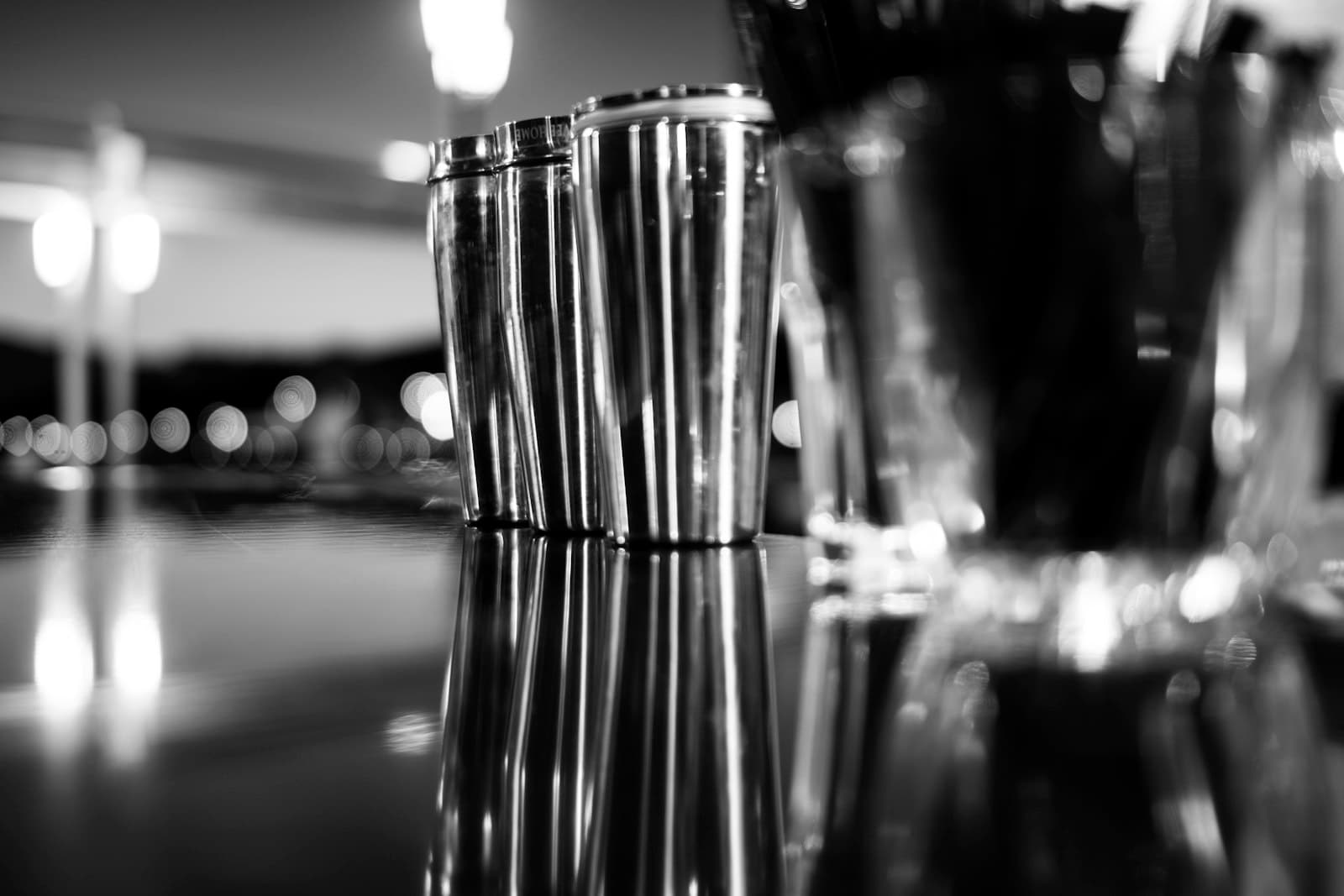 Tres cocteleras metálicas sobre la barra de un bar de noche, con luces desenfocadas al fondo, en blanco y negro
