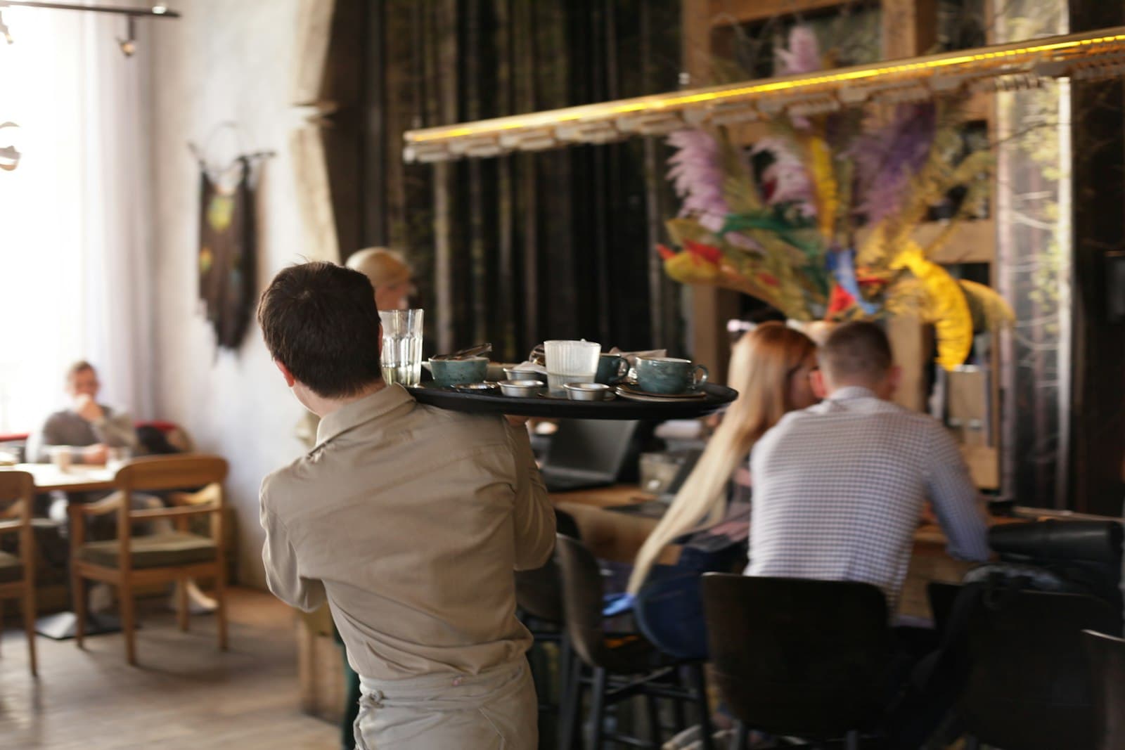 Camarero de espaldas cruzando una cafetería con una bandeja de tazas mientras los clientes charlan en las mesas