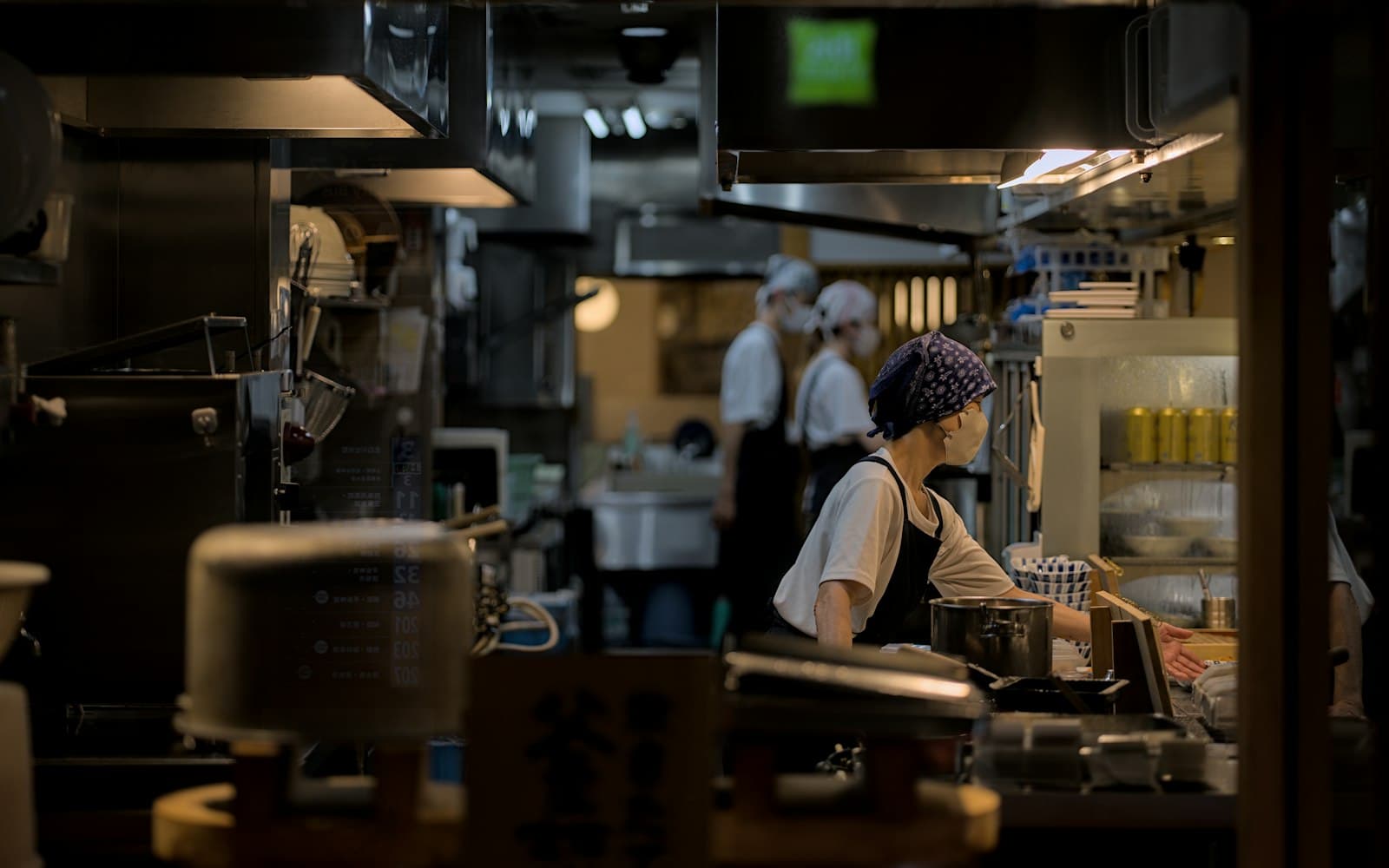 Una cocinera con pañuelo en la cabeza trabaja en el pase de una cocina, con dos compañeros al fondo