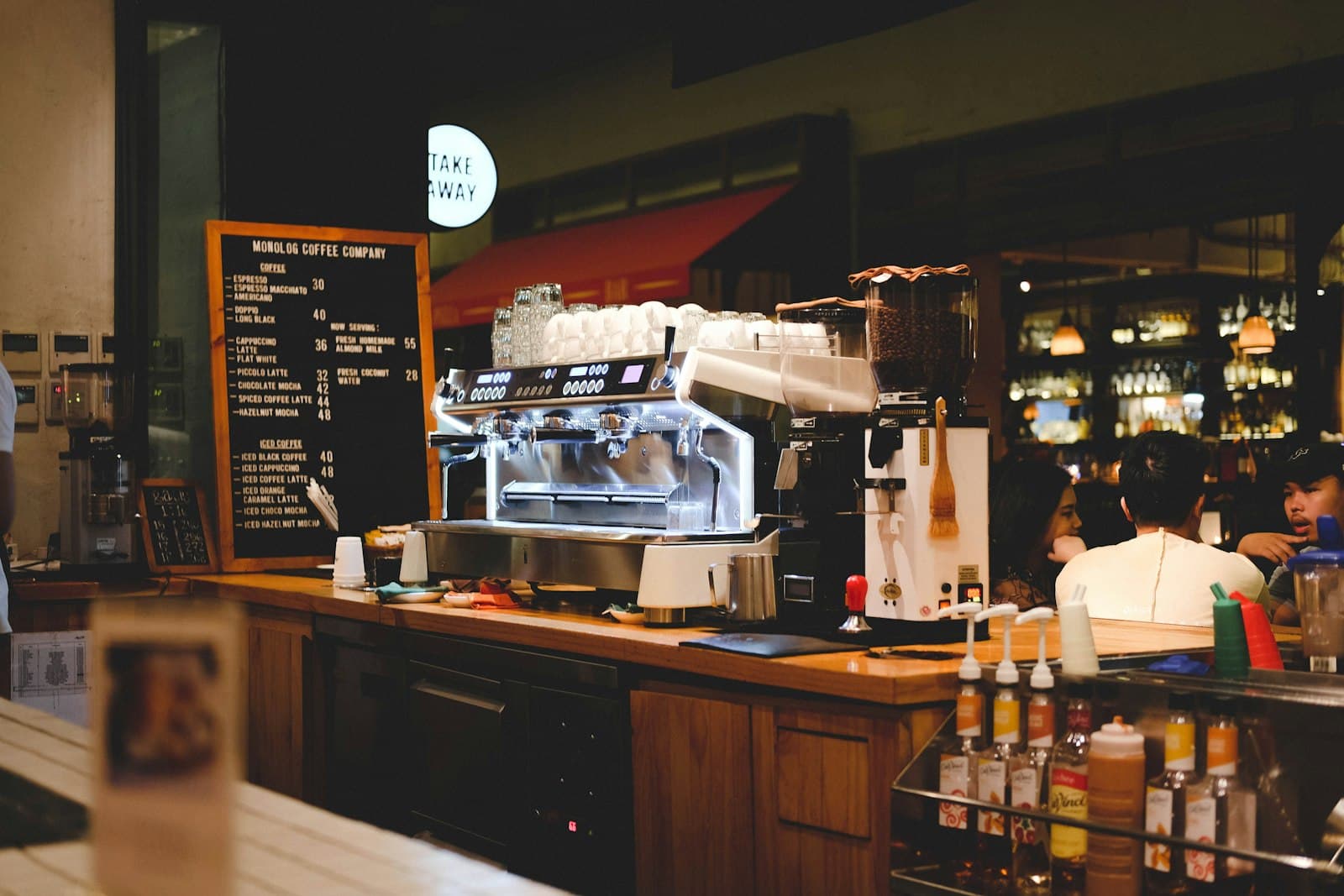 Barra de una cafetería con la cafetera, la pizarra de precios y clientes esperando al fondo