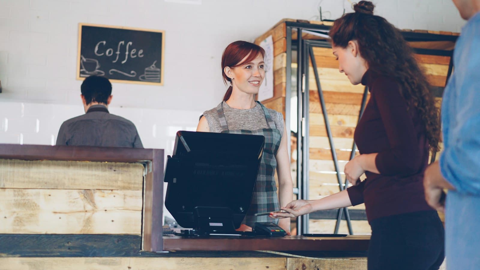 Una clienta paga en el mostrador de una cafetería mientras la camarera la atiende sonriendo detrás de la caja