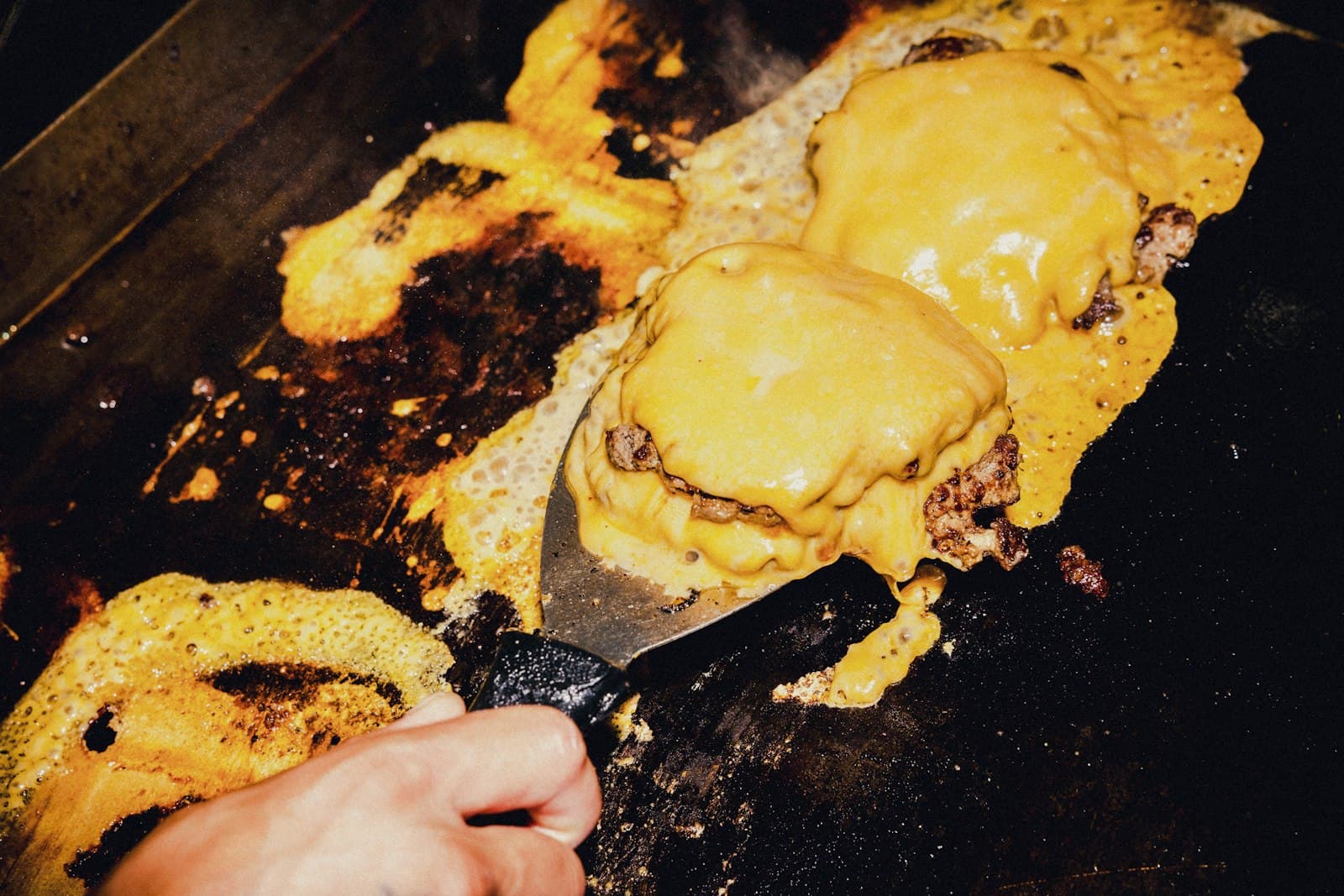 Mano con una espátula levantando hamburguesas con queso fundido en una plancha