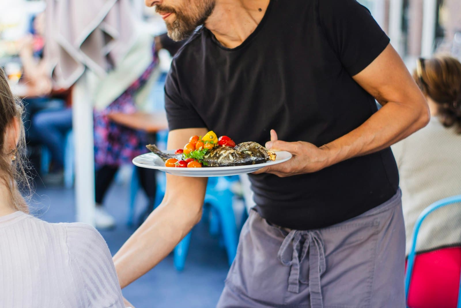 Camarero llevando un plato de pescado con tomates a una mesa en una terraza