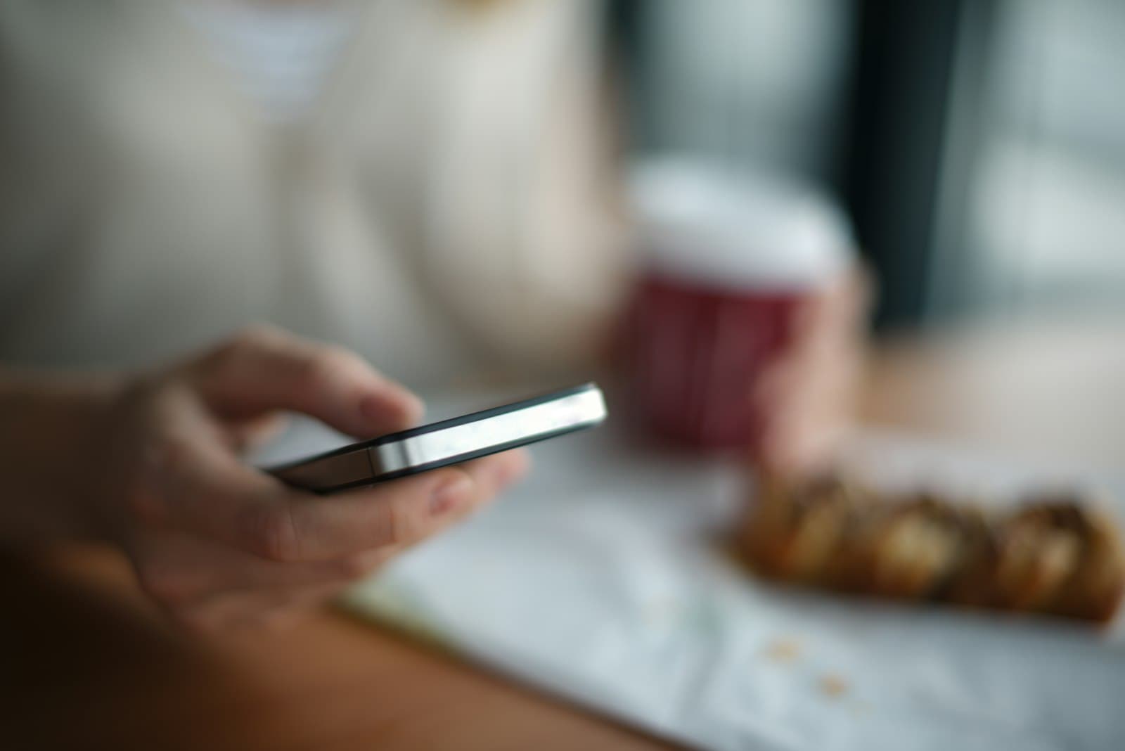 Mano sujetando un móvil en una cafetería, con un vaso de café y un bollo desenfocados al fondo