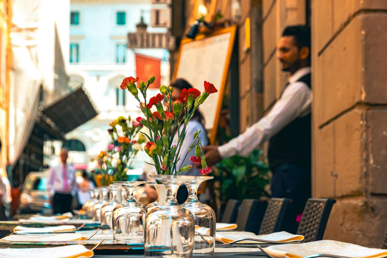Mesa de terraza preparada con copas y claveles, con un camarero al fondo junto a la puerta del restaurante