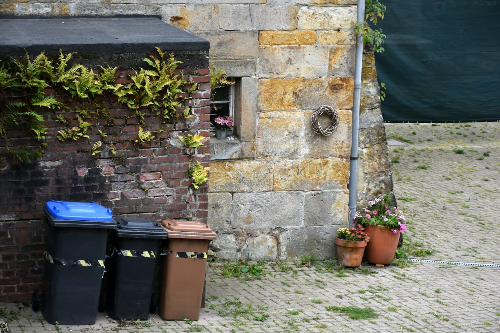 Contenedores de basura con tapas de colores junto a la pared de piedra de un edificio, con macetas de flores