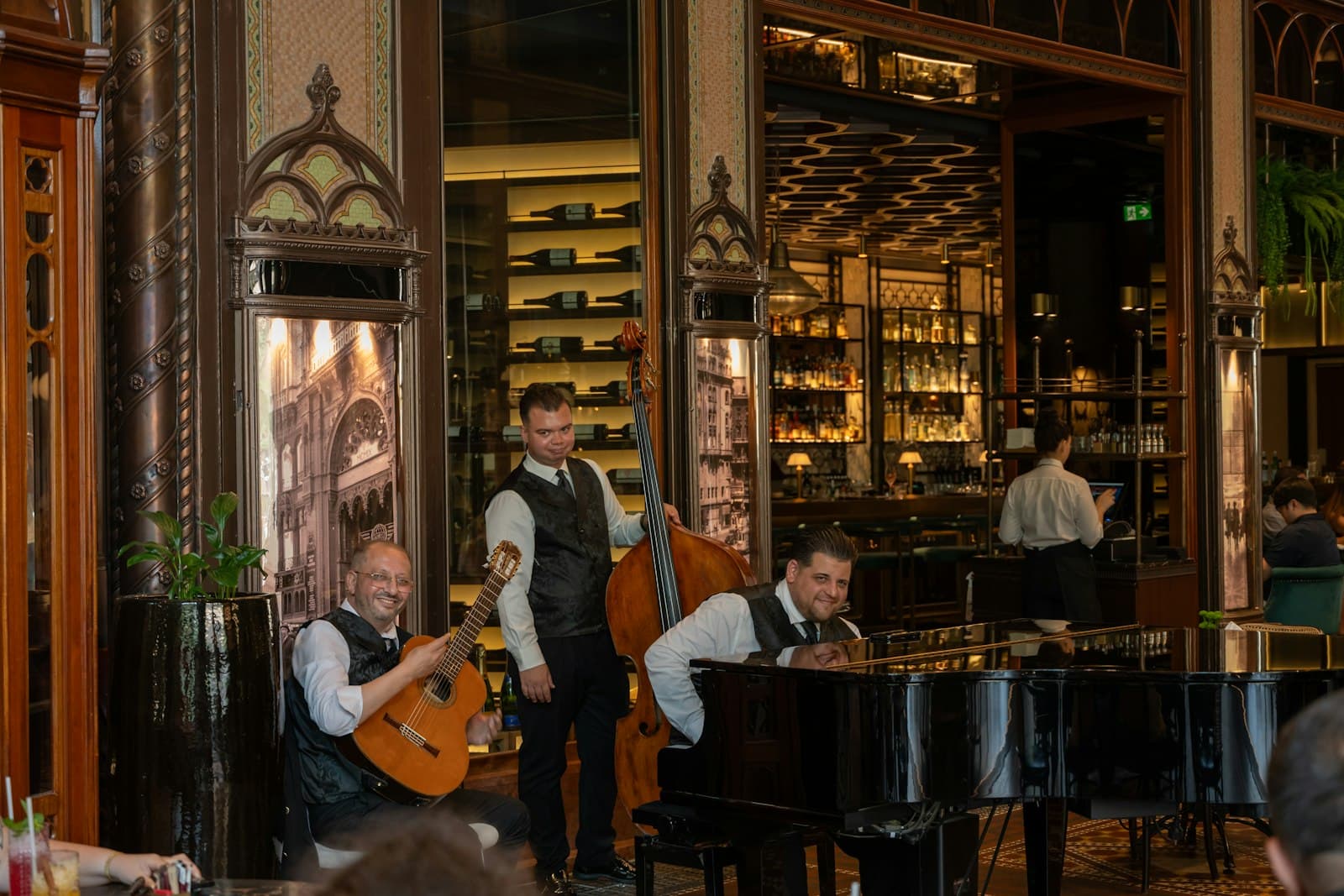 Tres músicos tocan guitarra, contrabajo y piano en directo en un restaurante, con la barra y las botellas al fondo