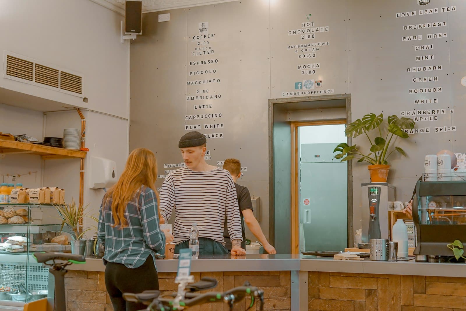 Una clienta pide en la barra de una cafetería mientras el camarero la atiende, con la carta de cafés escrita en la pared