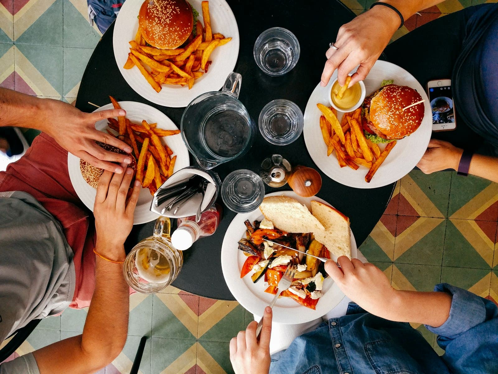 Cuatro personas comparten una mesa redonda con hamburguesas, patatas fritas, verduras asadas y agua, vista desde arriba