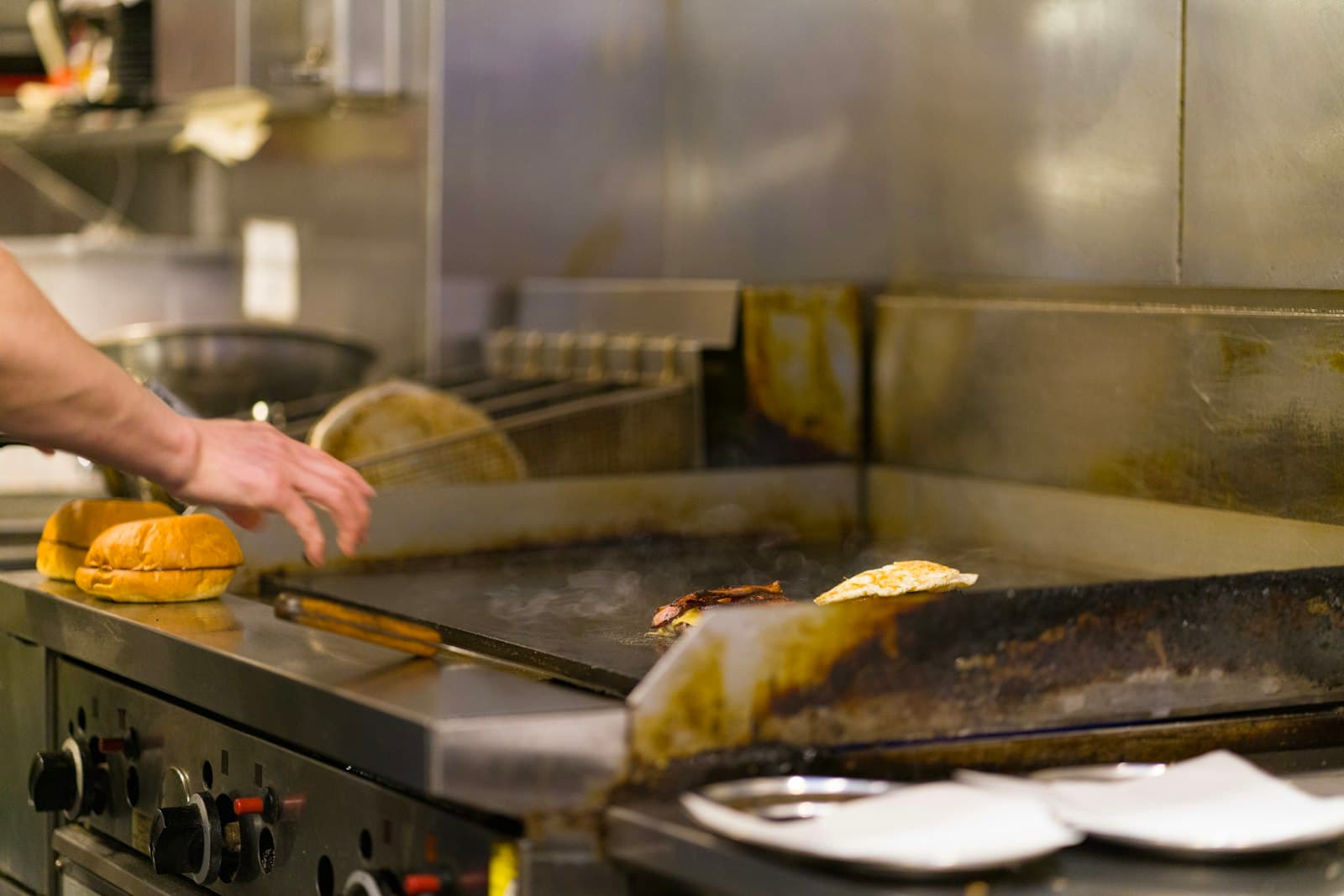 Mano de un cocinero junto a una plancha profesional con carnes de hamburguesa y panes esperando al lado