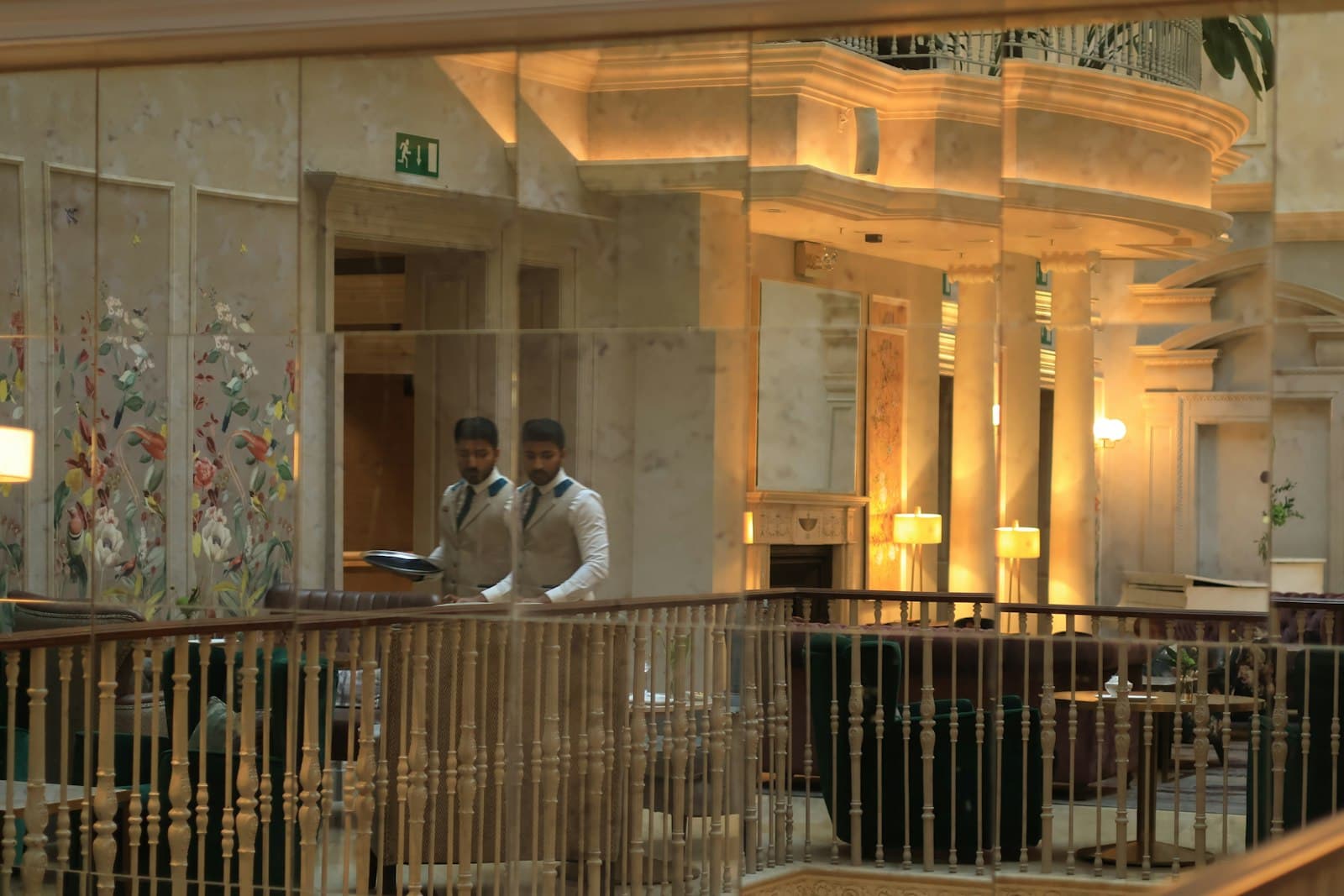 Un camarero con chaleco y una bandeja, reflejado en los espejos del salón de un hotel, con columnas, lámparas y butacas