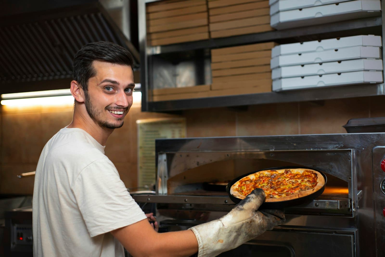 Pizzero sacando una pizza de un horno eléctrico de solera, con cajas de pizza para llevar apiladas detrás