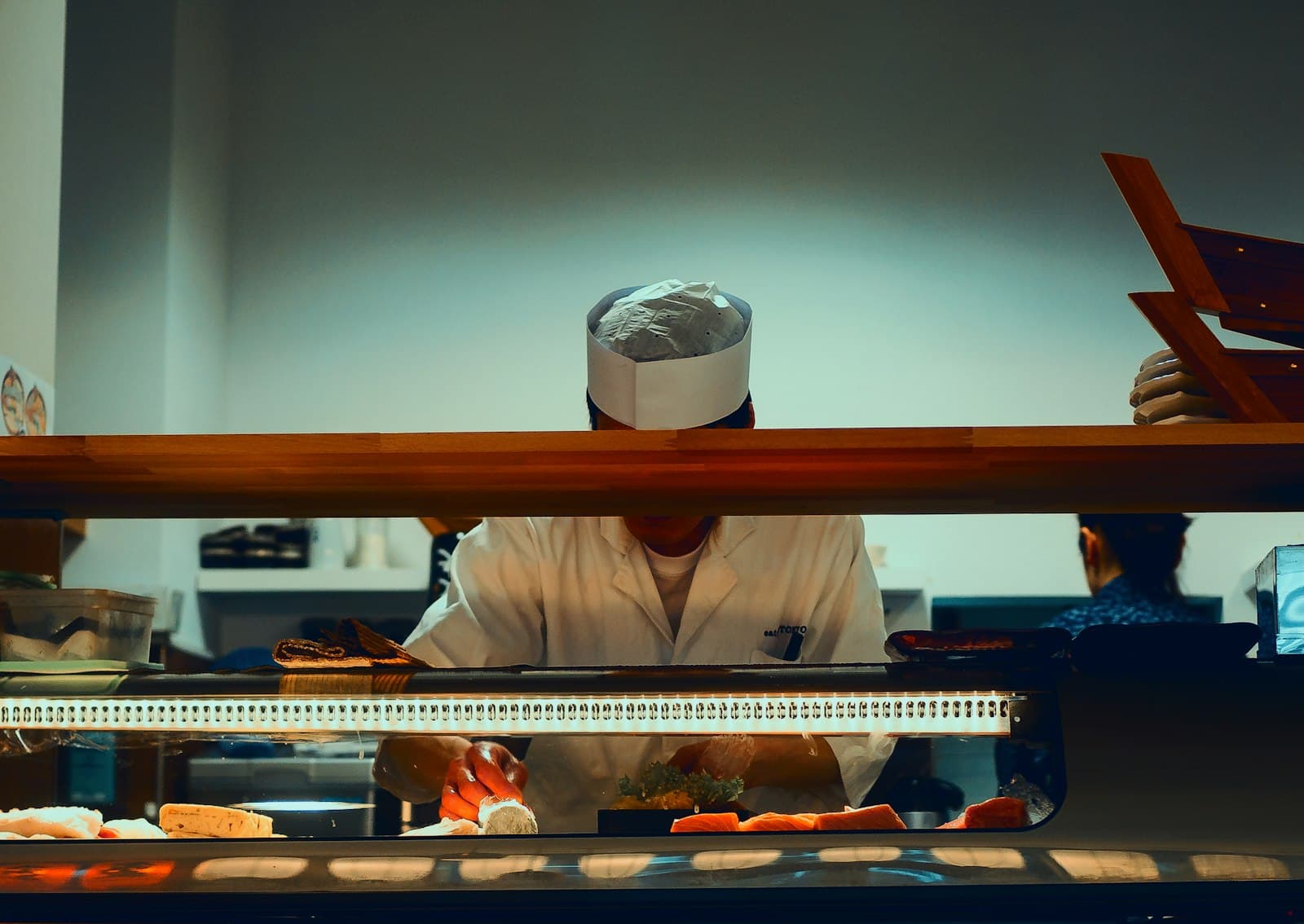 Cocinero de sushi preparando piezas detrás de la vitrina refrigerada de la barra, con lomos de salmón y atún a la vista