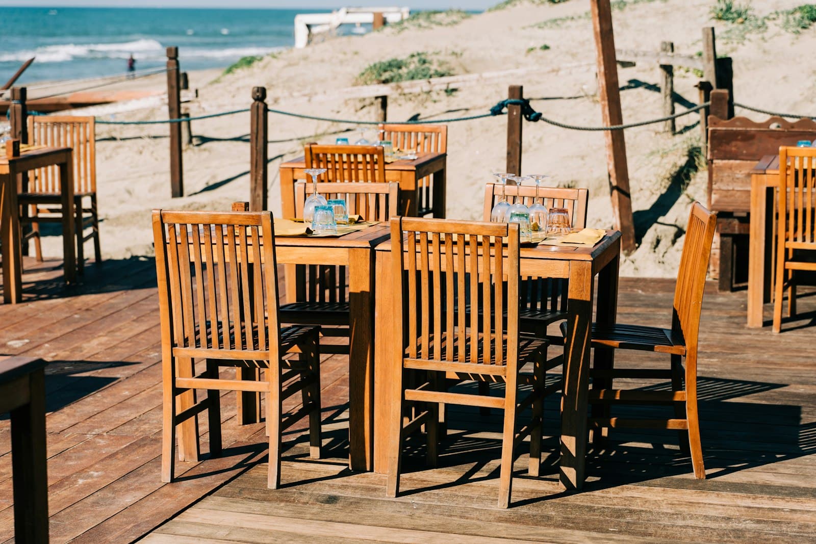 Mesas de madera montadas con copas en la tarima de un restaurante de playa, con el mar al fondo