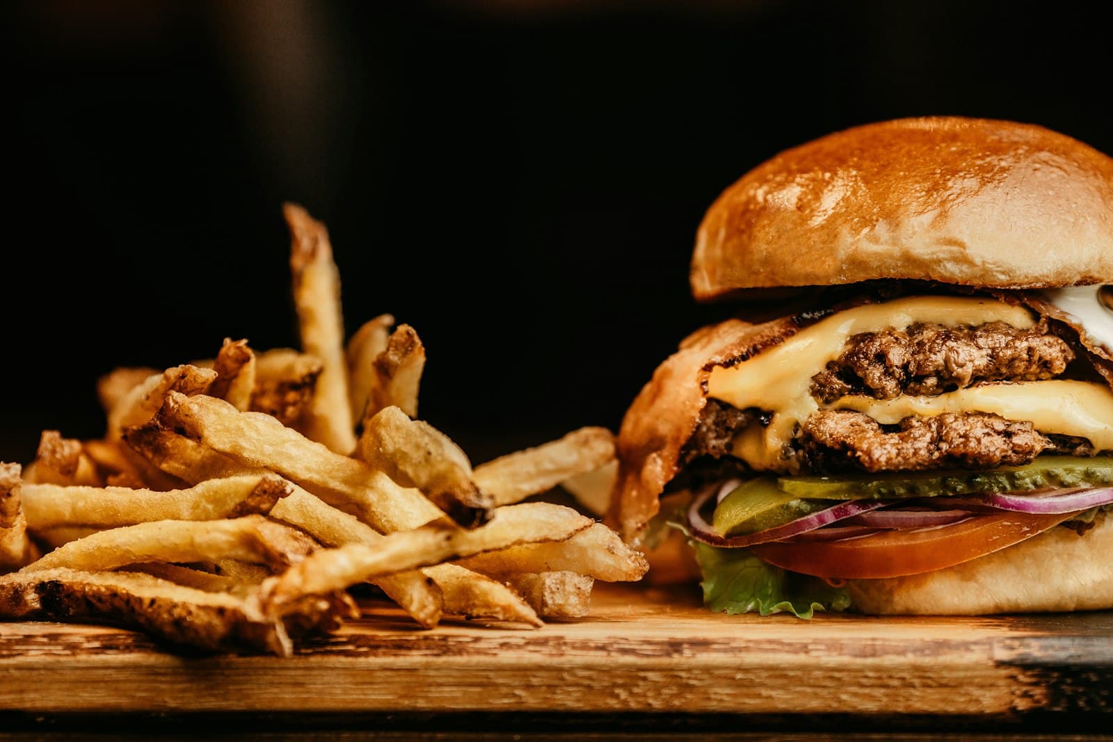 Hamburguesa doble con queso, cebolla roja y pepinillos junto a unas patatas fritas sobre una tabla de madera