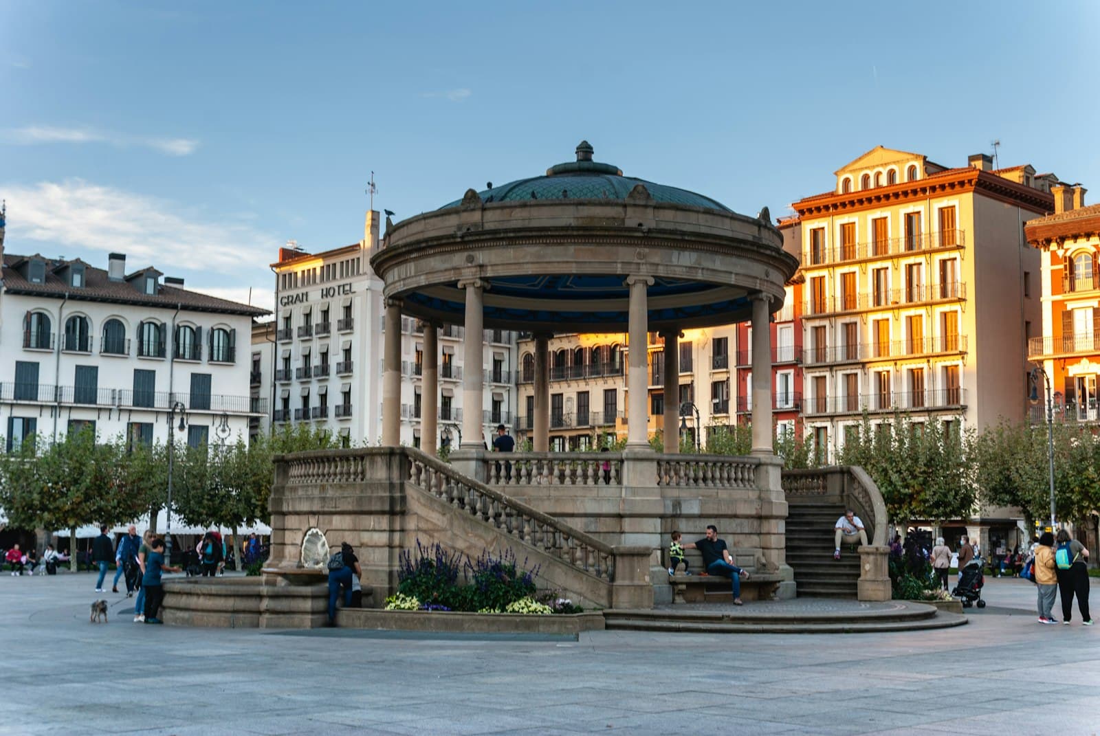 Quiosco de la plaza del Castillo de Pamplona al atardecer, con gente sentada en sus escaleras y los edificios de la plaza al fondo