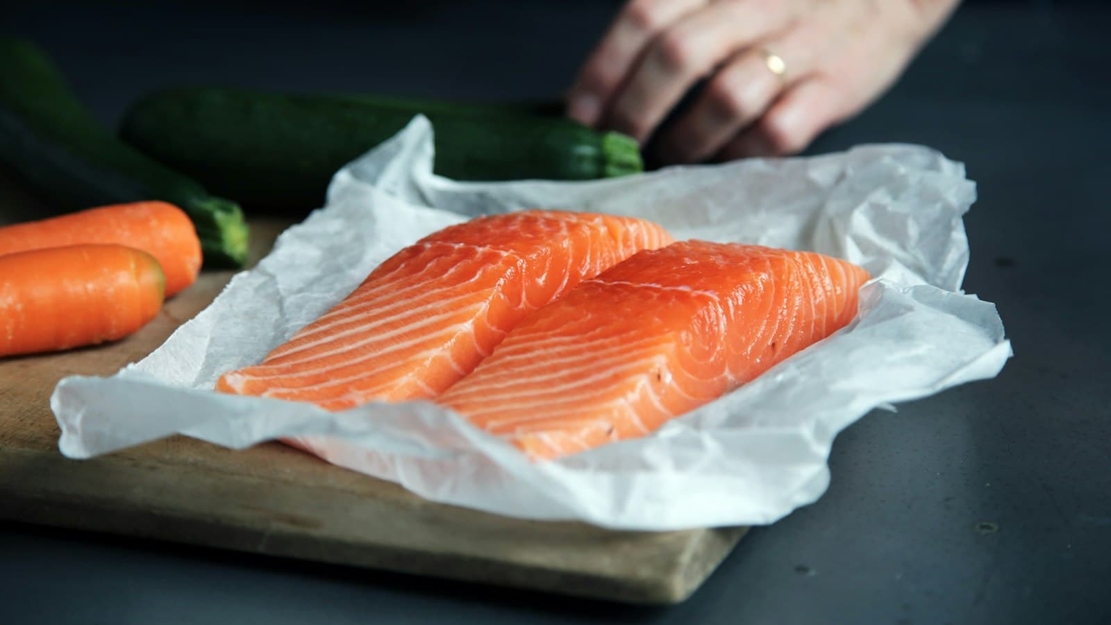 Dos lomos de salmón crudo sobre papel de cocina en una tabla de madera, con zanahorias y calabacines al fondo
