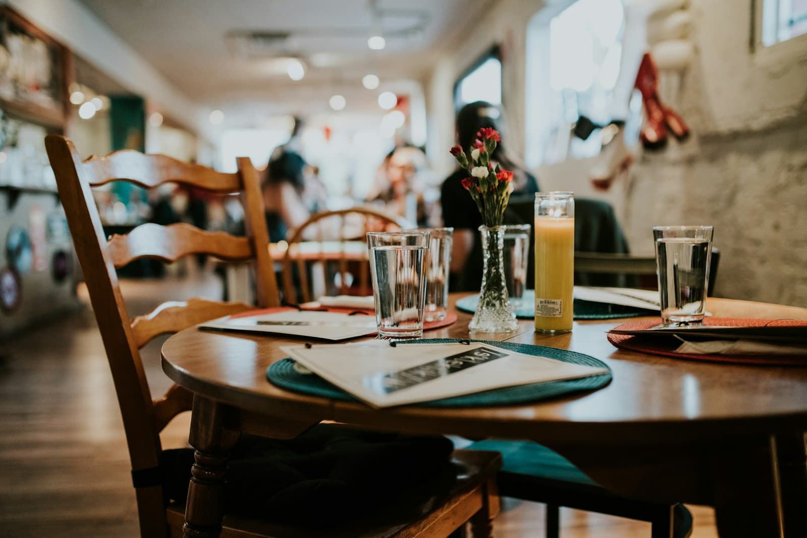 Mesa de restaurante con vasos de agua, zumo, flores y cartas, con clientes desenfocados al fondo