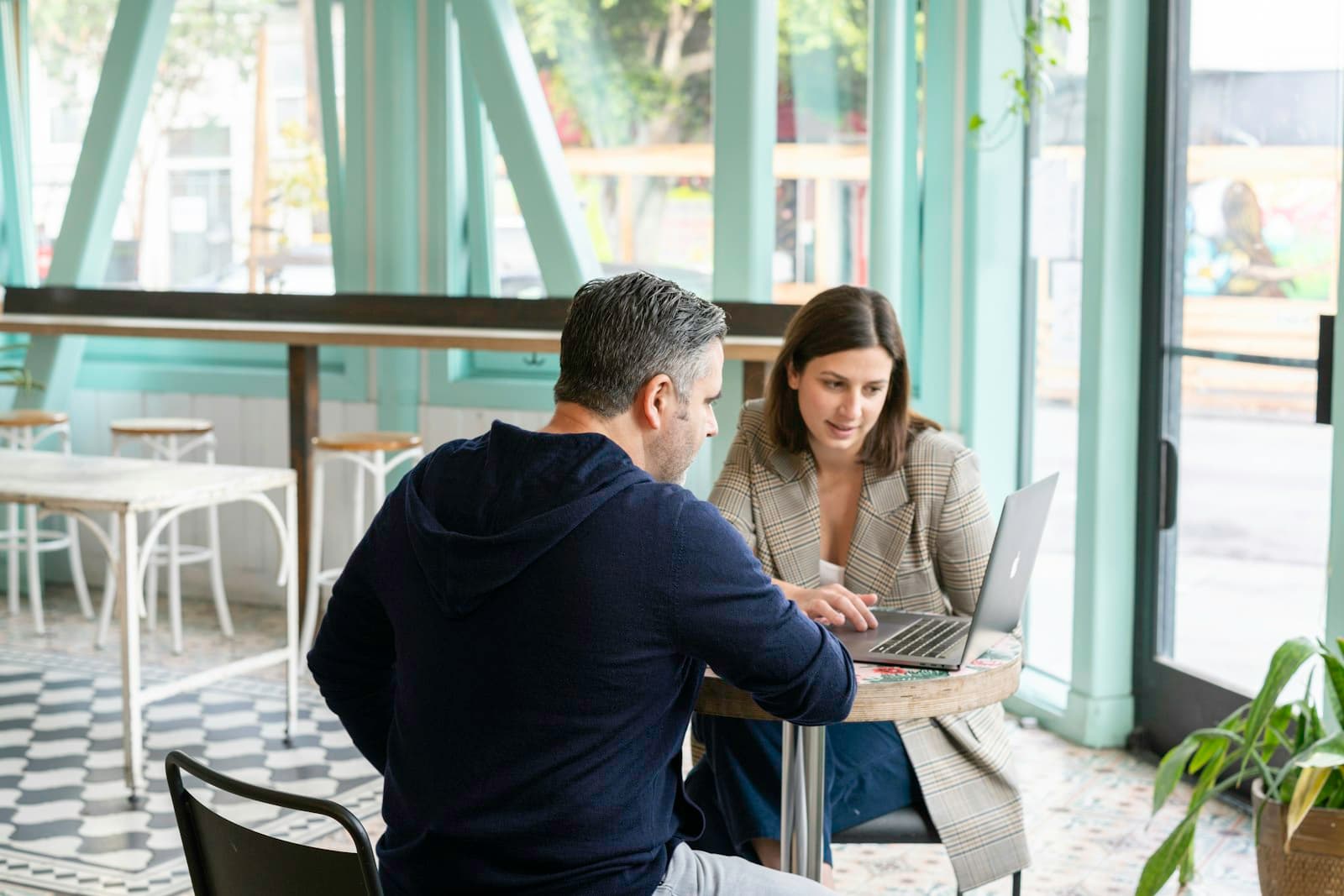 Dos personas revisan algo en un portátil en la mesa de una cafetería con grandes ventanales