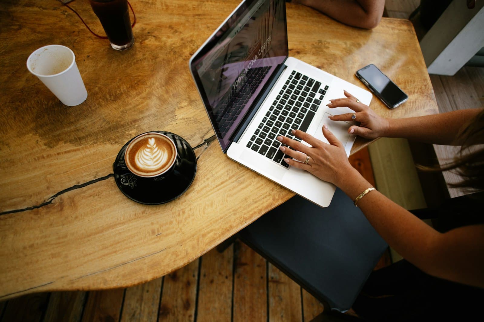 Persona escribiendo en un portátil sobre una mesa de madera de cafetería, junto a un café con leche
