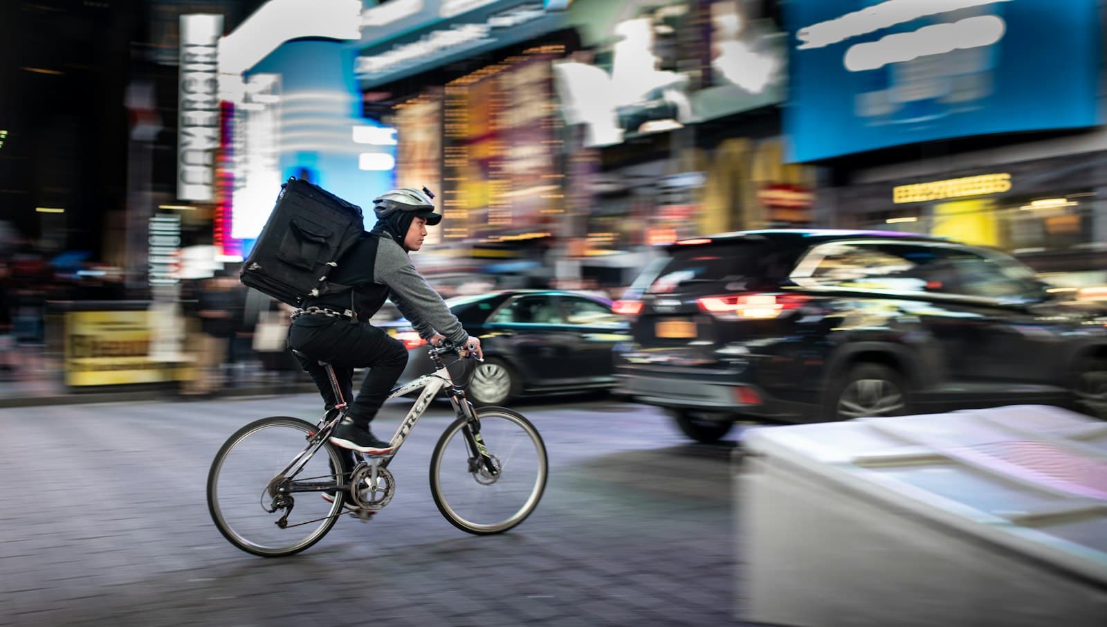 Repartidor en bicicleta con mochila térmica negra circulando entre coches por una avenida iluminada de noche