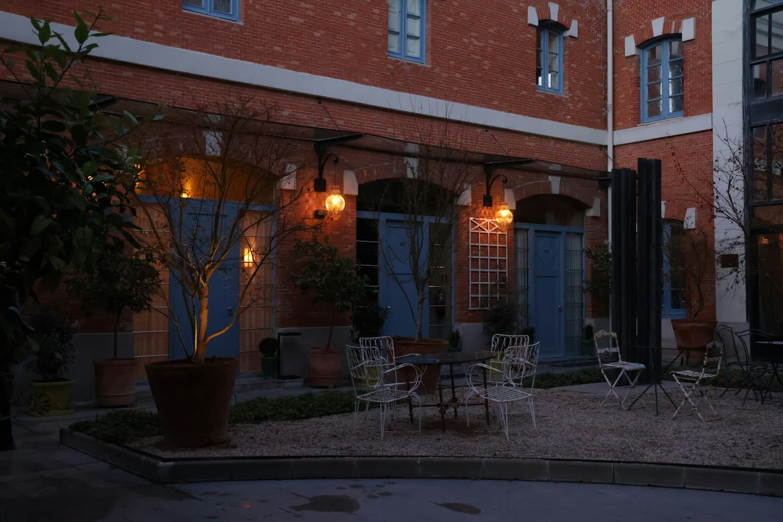 Terraza vacía en un patio de ladrillo de Madrid al anochecer, con mesas y sillas de forja y faroles encendidos