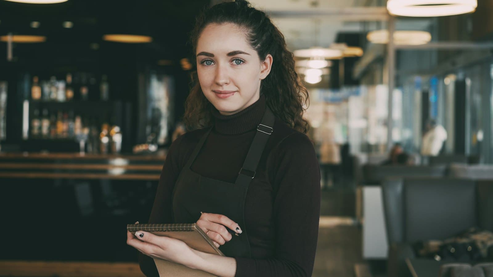 Camarera con delantal y una libreta en la mano, de pie delante de la barra de un restaurante