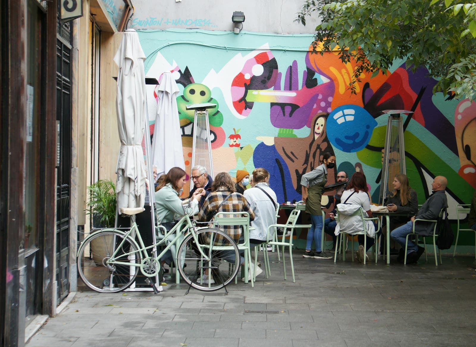 Terraza de un bar de Madrid con estufas de exterior, sombrillas cerradas y clientes sentados frente a un mural de colores