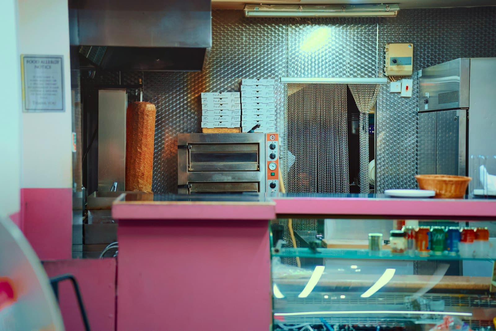 Mostrador rosa de un local de kebab y pizza con cajas de pizza apiladas y el horno al fondo
