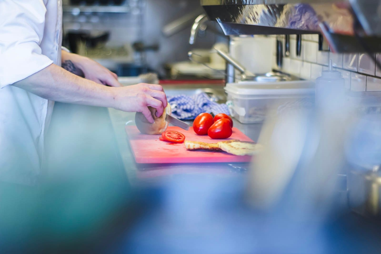 Cocinero cortando pan y tomates sobre una tabla roja en la encimera de una cocina profesional