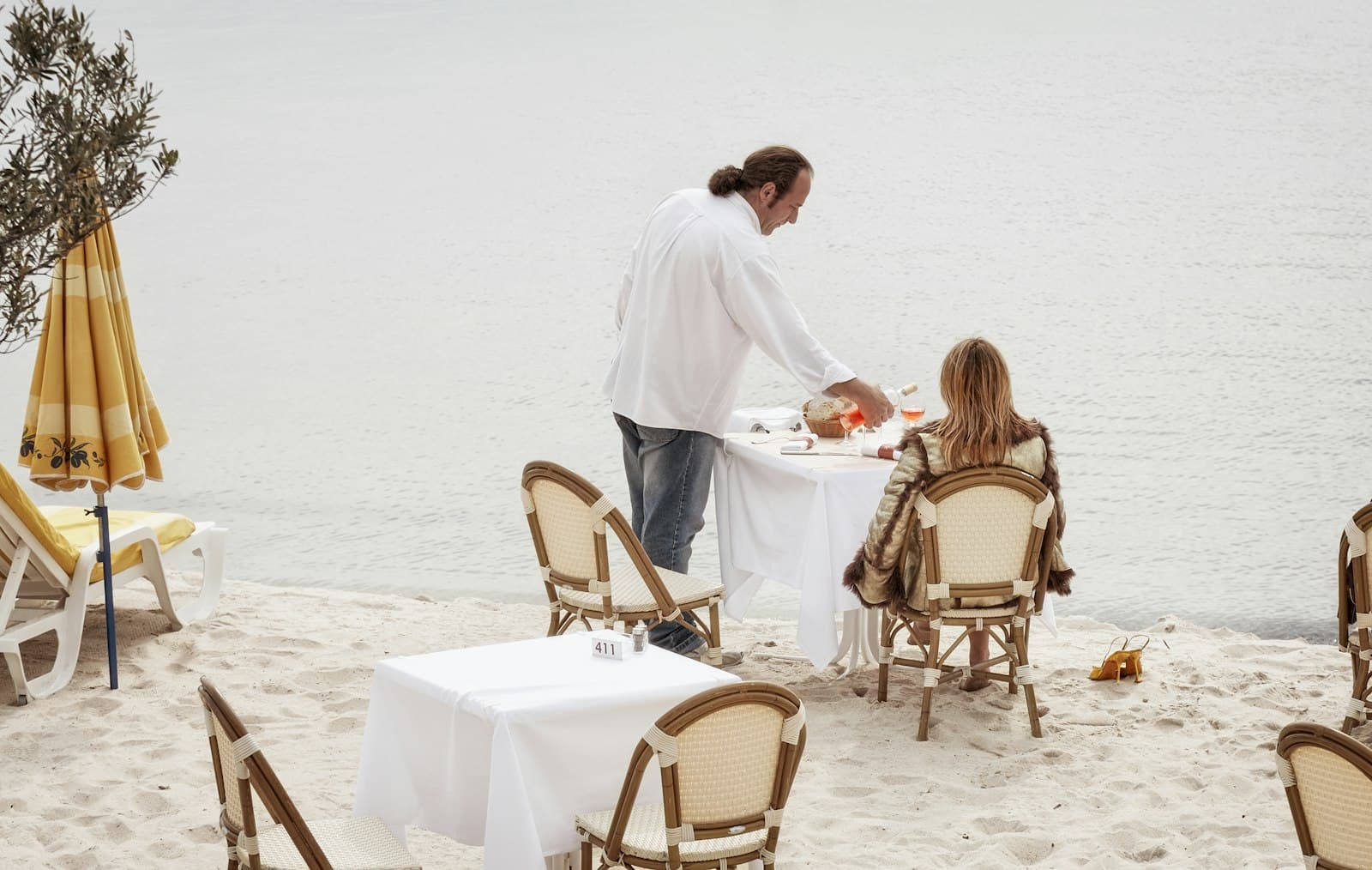Un camarero sirve vino a una clienta en una mesa con mantel blanco sobre la arena, junto a la orilla