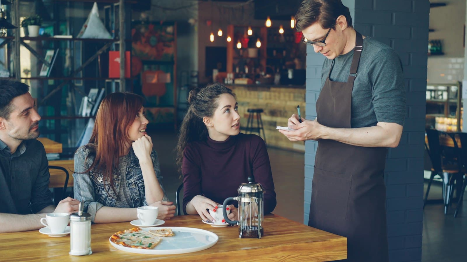 Camarero con delantal apuntando la comanda en una libreta junto a una mesa de clientes en una cafetería